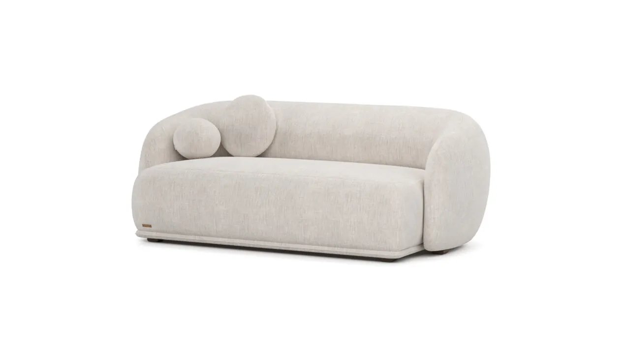 Kreminė Otis sofa su plačia vientisa sėdyne ir lenktu apgaubiančiu atlošu. Kairėje dvi apvalios pagalvėlės.